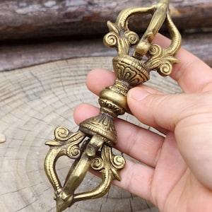May include: A close-up of a golden, ornate vajra, a ritual object with intricate scrollwork and a central handle. The vajra is held in a hand, showcasing its detailed design and aged appearance. The object is likely used for spiritual or ceremonial purposes.