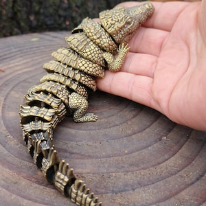 May include: A detailed brass articulated crocodile figurine. The sculpture features a textured, segmented body, legs, and head. The crocodile is a golden-brown colour and appears to be made of metal. The figurine is resting on a wooden surface.