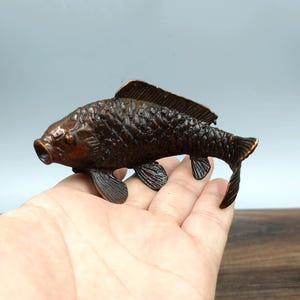 Puede incluir: Una escultura de carpa de color bronce con escamas, aletas y cola detalladas. El pez se representa con la boca abierta, y la escultura se sostiene en una mano. La carpa es un símbolo de buena fortuna y prosperidad.