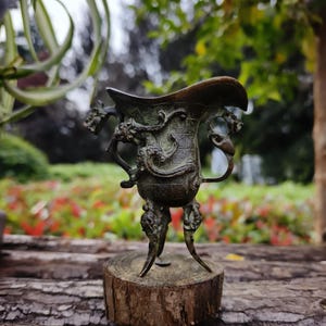 May include: A bronze-colored, ornate vessel with dragon motifs and handles. The vessel has a flared rim and is supported by three legs. It is mounted on a small, round wooden base, with a blurred background of greenery.