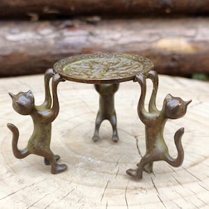 Könnte beinhalten: Ein dekorativer Tisch in Bronzefarbe, der von drei Katzenfiguren getragen wird. Die runde Tischplatte hat ein geprägtes Design. Die Katzen befinden sich in einer Hebehaltung und schaffen ein skurriles und künstlerisches Stück.