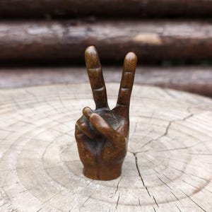 May include: A bronze-colored sculpture of a hand making the peace sign. The fingers are raised in a V shape, with the palm facing forward. The sculpture is set on a light-colored, textured wooden surface.