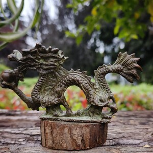 May include: A bronze dragon sculpture with intricate details, holding a sphere. The dragon is mounted on a small, round wooden base. The sculpture has a weathered, antique appearance, with a green patina.