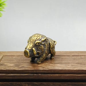 Puede incluir: Figurilla de latón detallada de un jabalí, que muestra texturas intrincadas y un acabado envejecido. La escultura presenta colmillos prominentes y una frente arrugada, capturando la forma robusta del animal. El jabalí está posicionado sobre una superficie de madera.