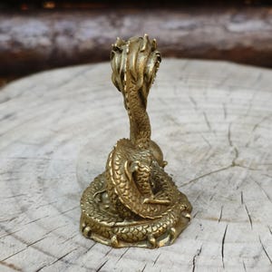 May include: A detailed brass dragon figurine. The dragon is coiled with its head raised, showcasing intricate scales and a textured surface. The figurine has a vintage aesthetic and is set against a natural wood background.