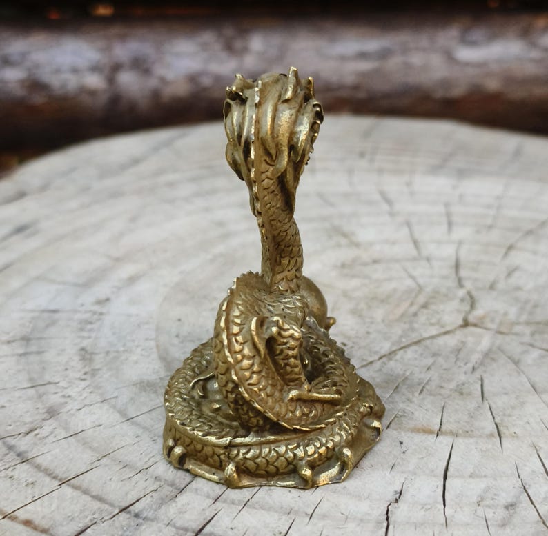 May include: A detailed brass dragon figurine. The dragon is coiled with its head raised, showcasing intricate scales and a textured surface. The figurine has a vintage aesthetic and is set against a natural wood background.