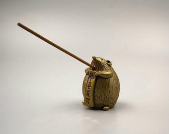 May include: A bronze-colored incense holder shaped like a small, round animal. The animal holds a banner with Chinese characters and has an incense stick inserted into its mouth. The incense stick is a light brown color.