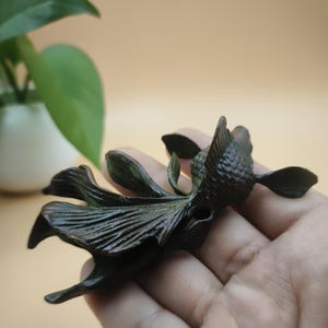 May include: A dark bronze-colored sculpture of a stylized flower or aquatic creature. The sculpture has detailed petal-like structures and a textured surface. The piece is held in a hand, with a blurred background of a plant in a white pot.