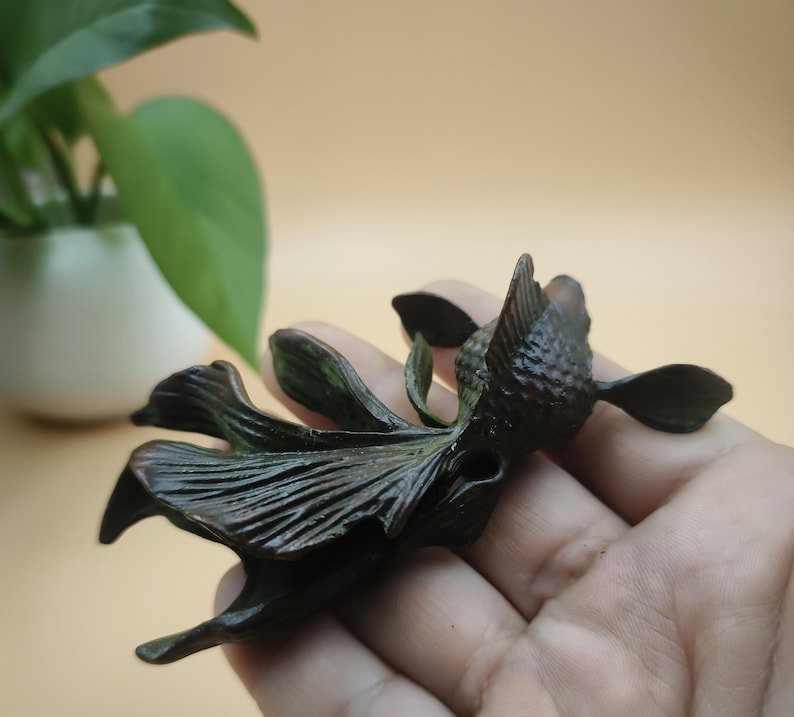 May include: A dark bronze-colored sculpture of a stylized flower or aquatic creature. The sculpture has detailed petal-like structures and a textured surface. The piece is held in a hand, with a blurred background of a plant in a white pot.