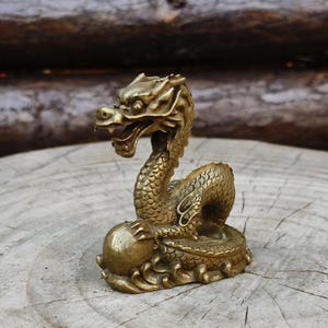 May include: A detailed brass dragon figurine. The dragon is posed with its head raised and mouth open, holding a sphere. The sculpture has intricate scales and details, with a weathered, antique finish. The dragon is approximately 4 inches tall.