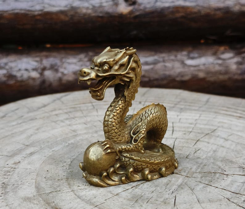 May include: A detailed brass dragon figurine. The dragon is posed with its head raised and mouth open, holding a sphere. The sculpture has intricate scales and details, with a weathered, antique finish. The dragon is approximately 4 inches tall.