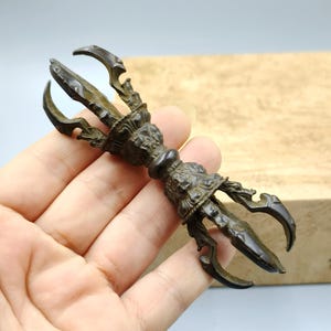 May include: A bronze-coloured Vajra, a ritual object with three prongs on each end, held in a hand. The central part features intricate carvings and a bulbous centre. The object is likely used in religious or spiritual practices.