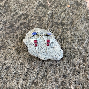 May include: A pair of silver-toned earrings featuring a triangular frame with two rectangular red stones and a single oval blue stone at the top. The earrings have a metallic finish and are designed with a hook closure for attachment to the ear.