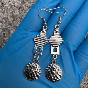 May include: A pair of silver-toned dangle earrings featuring a tiered, geometric top section connected to a circular, hammered-texture pendant. The earrings are suspended from standard silver-colored metal ear hooks with a polished finish.