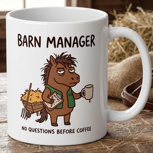 May include: A white ceramic mug featuring a cartoon brown horse wearing a green vest, holding a clipboard labeled DUTIES and a coffee mug. The text BARN MANAGER appears above the horse, and NO QUESTIONS BEFORE COFFEE is printed below the illustration.