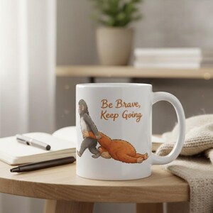 May include: White ceramic mug with a whimsical illustration of a monkey carrying an orangutan, with the text "Be Brave, Keep Going" in brown. The mug has a curved handle and is set on a wooden table.