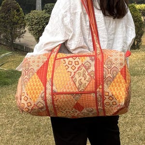 Può includere: Una grande borsa tote rettangolare con un design patchwork nei toni dell'arancione, giallo e rosso. La borsa ha una tasca rettangolare sul davanti e cinghie rosse. La borsa viene portata a spalla.
