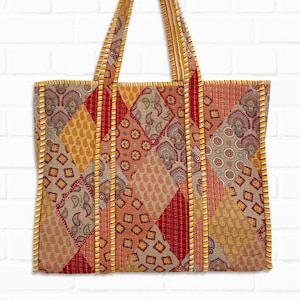 Op de afbeelding: Een gewatteerde tote bag met een patchwork design. De tas heeft een verscheidenheid aan warme kleuren, waaronder rood, oranje, geel en bruin. De tas heeft twee handvatten en is gemaakt van stof. De tas hangt tegen een witte bakstenen muur.