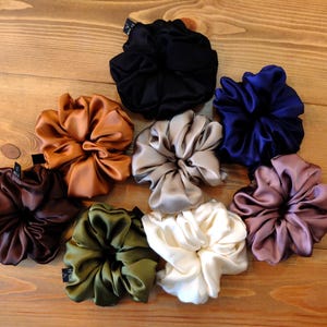 Op de afbeelding: Een verzameling satijnen scrunchies in diverse kleuren, waaronder zwart, marineblauw, bruin, goud, olijfgroen, crème, taupe en mauve. De scrunchies liggen op een houten oppervlak.