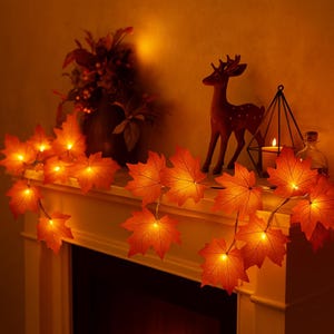 Può includere: Una scena calda e accogliente con una ghirlanda di luci a forma di foglie d'acero arancioni drappeggiata su un caminetto. Una piccola statuina di cervo e una candela in un supporto geometrico contribuiscono all'atmosfera autunnale. È visibile anche un vaso di fiori secchi.
