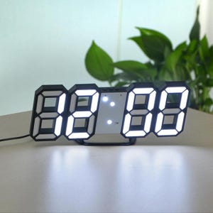Può includere: Un moderno orologio digitale con cornice nera e numeri LED bianchi luminosi. L'orologio visualizza l'ora in stile 3D, con i numeri che sembrano fluttuare. L'orologio è su una superficie bianca, con fogliame verde sullo sfondo.