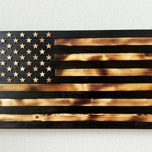 May include: Wooden American flag wall art. The flag features a distressed design with black and natural wood tones. The stars are individually cut and attached. The flag is rectangular and mounted on a white wall.