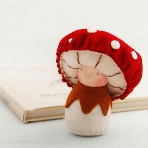 May include: A handmade mushroom plush toy with a red cap and white spots, a beige underside, and a brown stem. The mushroom is set in front of a closed book with a beige cover. The website address www.shroompers.com is visible.