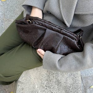 以下が含まれることがあります： クロコダイル柄のダークブラウンのテクスチャードクラッチバッグ。グレーのコートとオリーブグリーンのパンツを着た人が持っています。バッグはギャザーデザインの長方形です。