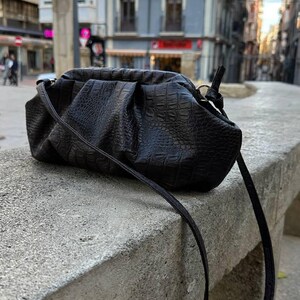 以下が含まれることがあります： 黒色のクラッチバッグ。クロコダイルのような質感の模様が施されています。細いストラップが付いており、コンクリートの縁に置かれています。背景には、建物が並ぶ街並みが写っています。