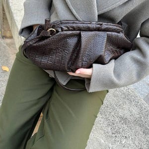 以下が含まれることがあります： ダークブラウンの、質感のある革製ハンドバッグ。ギャザーのデザインが施されています。ゴールドカラーのリングとショルダーストラップが付いています。グレーのコートとオリーブグリーンのパンツを着用しています。