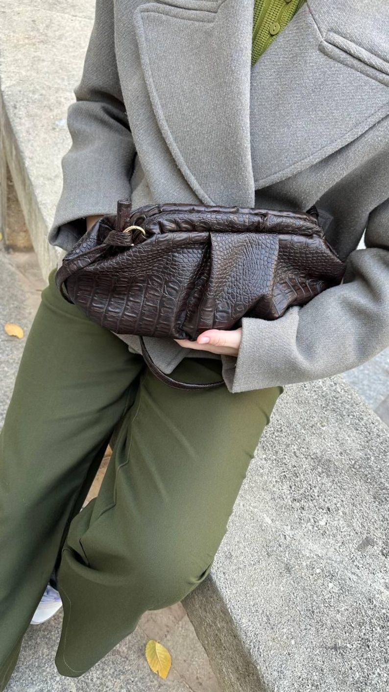 以下が含まれることがあります： ダークブラウンの、質感のある革製ハンドバッグ。ギャザーのデザインが施されています。ゴールドカラーのリングとショルダーストラップが付いています。グレーのコートとオリーブグリーンのパンツを着用しています。