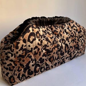 Op de afbeelding: Een gewatteerde clutch met een luipaardprint in bruin- en zwarttinten. De bovenkant van de clutch is gerimpeld met een donkerbruine stof. De tas heeft een zacht, opgeblazen uiterlijk.