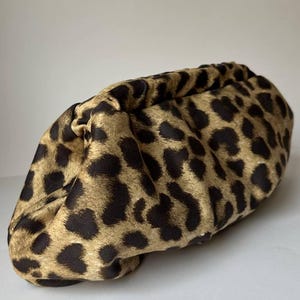 May include: A clutch bag with a leopard print pattern. The bag features a tan background with black spots. The bag is made of a soft, gathered material and has a rounded shape.