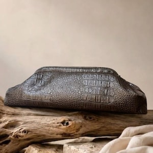 May include: A dark brown clutch bag featuring a textured, embossed crocodile skin pattern. The bag has a structured, elongated shape with a curved top edge and a smooth, consistent finish across its entire surface, showcasing a detailed scale design.