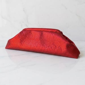 Puede incluir: Un bolso de mano de color rojo vibrante con una estructura alargada y un sutil patrón geométrico repetitivo en su superficie texturizada. El accesorio presenta un diseño elegante y cónico con un cierre de bisagra y un acabado metálico.