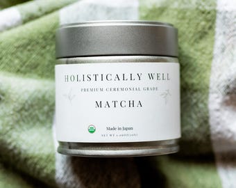 May include: A silver-lidded tin of "Holistically Well" Matcha, a premium ceremonial grade green tea powder. The label includes the text "Made in Japan" and the net weight of 1.06 oz (30g). The tin rests on a green and white checkered cloth.