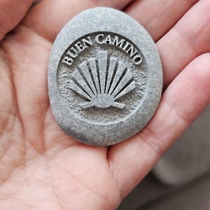 May include: A smooth, oval-shaped grey stone featuring an engraved scallop shell design in the centre. The text "BUEN CAMINO" is carved in a circular arc above the shell motif. The stone has a textured, matte finish and a natural, speckled appearance.