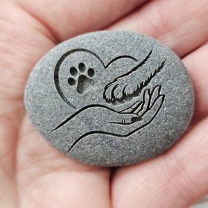 Peut inclure: Une pierre grise lisse de forme ovale avec un motif gravé au laser. La gravure représente une main humaine berçant une patte, avec une forme de cœur entourant l'empreinte de patte. La pierre présente une texture naturelle et mate.
