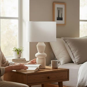 May include: A white table lamp with a cylindrical lampshade sits on a wooden bedside table. The lamp has a textured, rounded base. A person is reading a book next to the lamp. A small plant in a white pot and a mug are also on the table.