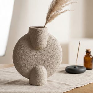 May include: A textured, beige ceramic vase with a circular body and two cylindrical openings. The vase holds dried ornamental grass. A small, dark gray incense holder and a brown glass bottle with a cork stopper are also present.