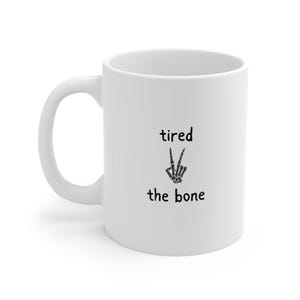 Pode incluir: Caneca de cerâmica branca com a inscrição preta "tired the bone" e um sinal de paz feito de ossos. A caneca tem uma alça curva e um acabamento liso e brilhante.