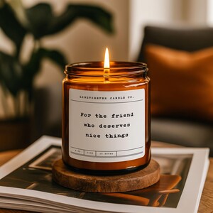 May include: A lit candle in an amber glass jar with a white label featuring the text Lightkeeper Candle Co. and For the friend who deserves nice things. The candle sits on a circular wooden base, showcasing a single glowing flame at the top.