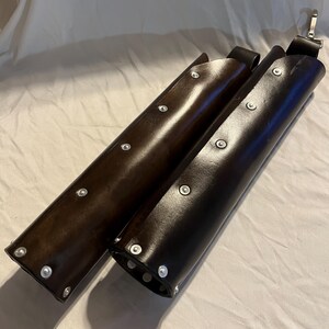 Puede incluir: Dos brazales de cuero marr&oacute;n. Los brazales son cil&iacute;ndricos, con remaches plateados en los bordes. Cada brazal tiene una correa negra con una hebilla plateada. Probablemente sean para tiro con arco o recreaci&oacute;n hist&oacute;rica.