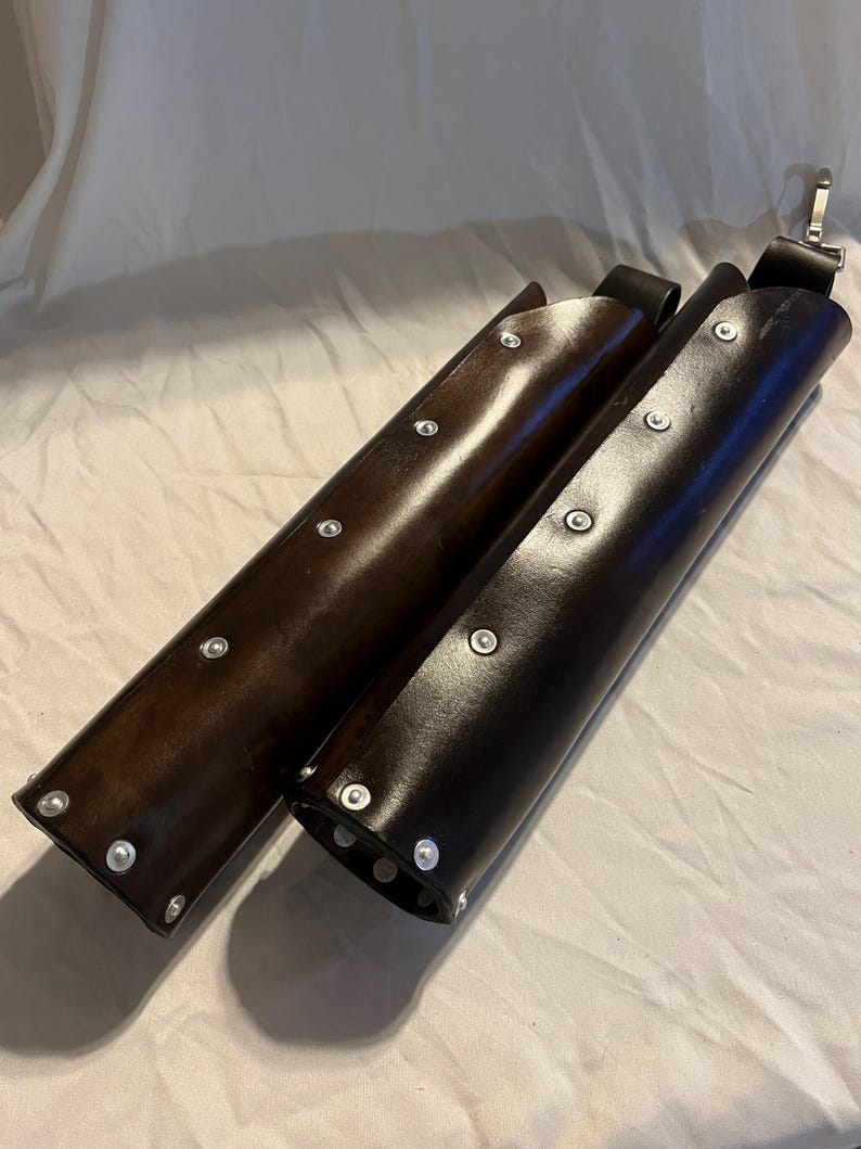 Puede incluir: Dos brazales de cuero marr&oacute;n. Los brazales son cil&iacute;ndricos, con remaches plateados en los bordes. Cada brazal tiene una correa negra con una hebilla plateada. Probablemente sean para tiro con arco o recreaci&oacute;n hist&oacute;rica.