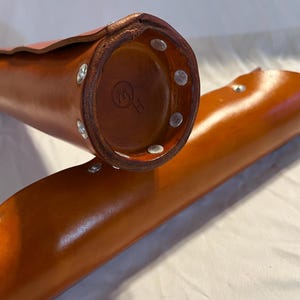 May include: Close-up of two cylindrical leather cases in a warm brown hue. The cases are secured with silver rivets. One case's base displays a circular emblem. The leather appears smooth and well-crafted, suggesting a focus on quality and durability.