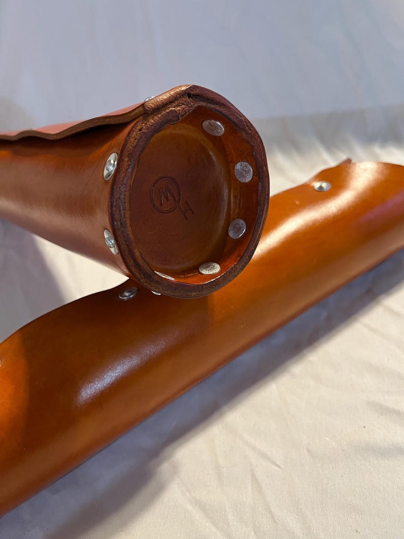 May include: Close-up of two cylindrical leather cases in a warm brown hue. The cases are secured with silver rivets. One case's base displays a circular emblem. The leather appears smooth and well-crafted, suggesting a focus on quality and durability.
