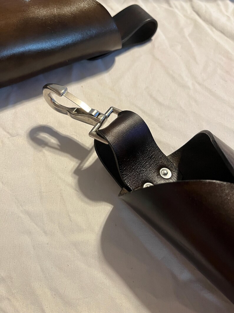 Puede incluir: Primer plano de una funda de cuero marr&oacute;n oscuro con un cierre de metal plateado. La funda tiene un lazo para un cintur&oacute;n y est&aacute; remachada. El fondo es una s&aacute;bana blanca.