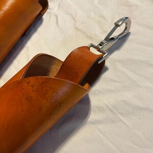 May include: Close-up of two orange-brown leather straps with a silver metal clasp. The leather has a textured appearance and is folded over. The clasp is attached to a loop of leather. The background is a white, wrinkled fabric.