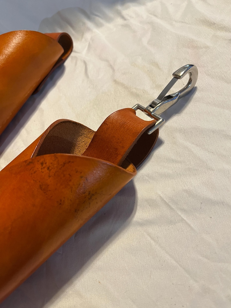 May include: Close-up of two orange-brown leather straps with a silver metal clasp. The leather has a textured appearance and is folded over. The clasp is attached to a loop of leather. The background is a white, wrinkled fabric.