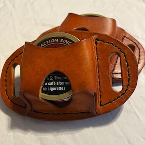 May include: Hand-stitched, brown leather tobacco pouch with black stitching. The pouch has a circular opening to display the tobacco tin inside. The text on the tin reads "ACTION SINCE" and "This product is a safe alternative to cigarettes."